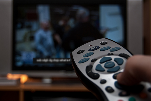 A person holds and points a remote control at a TV screen which has captions on the TV program’
